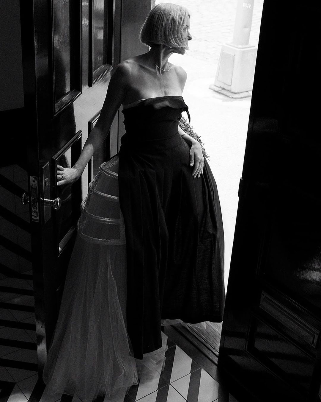Elegant woman in a strapless black dress and tulle petticoat stands in a doorway, looking off into the distance in a black and white portrait.