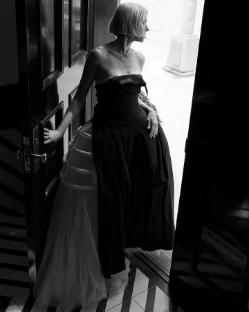 Elegant woman in a strapless black dress and tulle petticoat stands in a doorway, looking off into the distance in a black and white portrait.