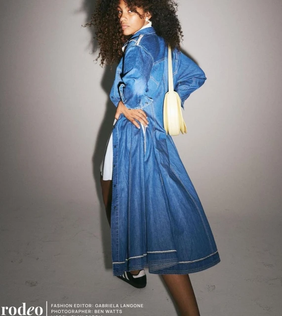 Fashion shot of a curly-haired woman wearing a long denim coat dress, handbag, and sneakers, posing against a grey backdrop.