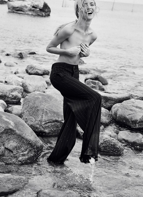 Monochrome portrait of a fair-skinned adult woman wading in water among rocks, laughing with hands covering her chest, wearing pants.