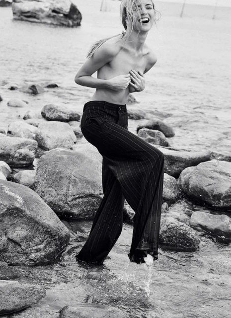 Monochrome portrait of a fair-skinned adult woman wading in water among rocks, laughing with hands covering her chest, wearing pants.