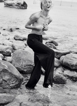 Monochrome portrait of a fair-skinned adult woman wading in water among rocks, laughing with hands covering her chest, wearing pants.