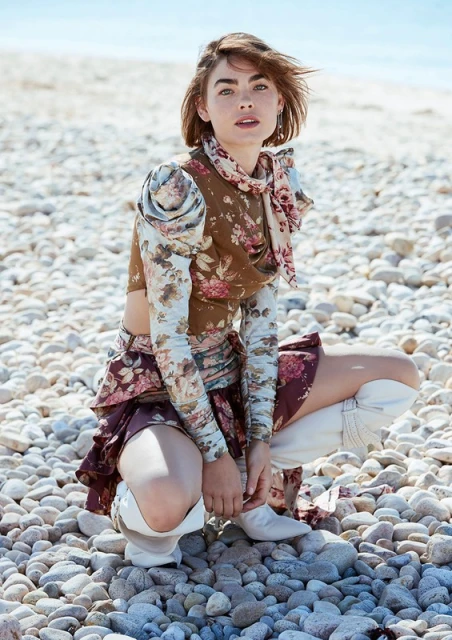 A woman in floral-print outfit crouches on a rocky beach in bright sunlight, looking at the viewer with a neutral expression.
