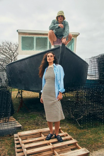 Two people pose with a boat: a woman in a striped dress stands on wooden pallets, and a man in a floral shirt crouches on the boat.