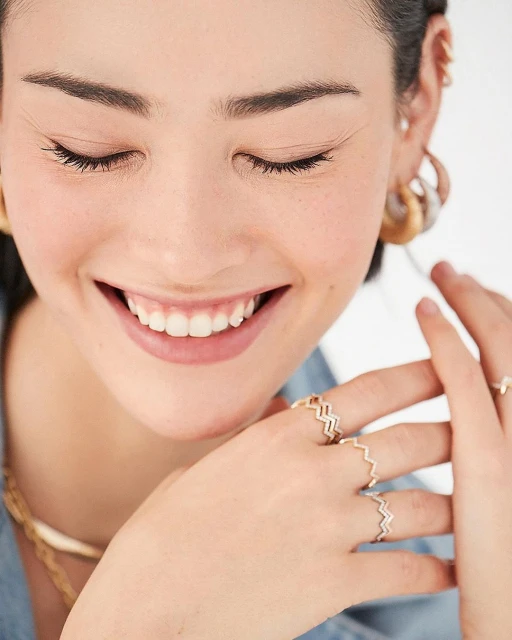 Smiling woman showcases gold zigzag rings; the jewelry is featured in a close-up shot against a bright background, highlighting the ring design.