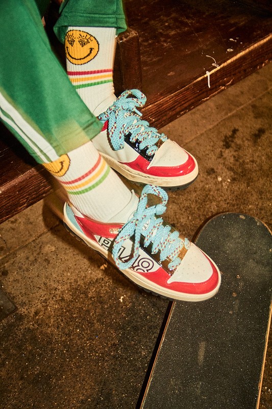 Close-up of white, red, and black low-top sneakers with light blue laces, worn with smiley-face socks, and placed on a skateboard.