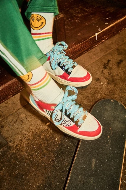 Close-up of white, red, and black low-top sneakers with light blue laces, worn with smiley-face socks, and placed on a skateboard.