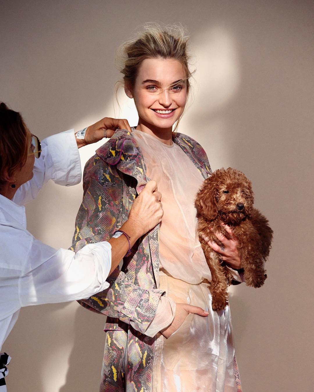 A young woman in an ornate patterned coat holds a red poodle as a stylist adjusts her collar against a neutral studio backdrop.