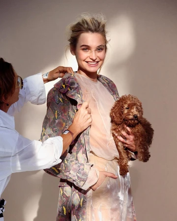 A young woman in an ornate patterned coat holds a red poodle as a stylist adjusts her collar against a neutral studio backdrop.