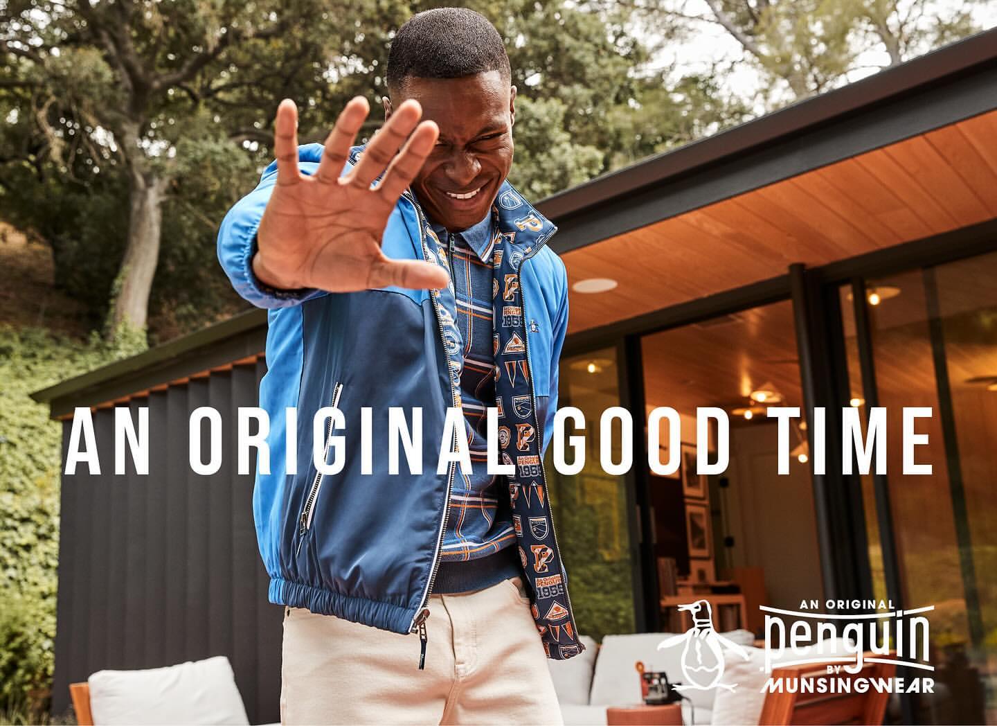 An African-American man smiles and puts his hand up in front of a building, wearing a blue jacket and khaki pants for a fashion advertisement.