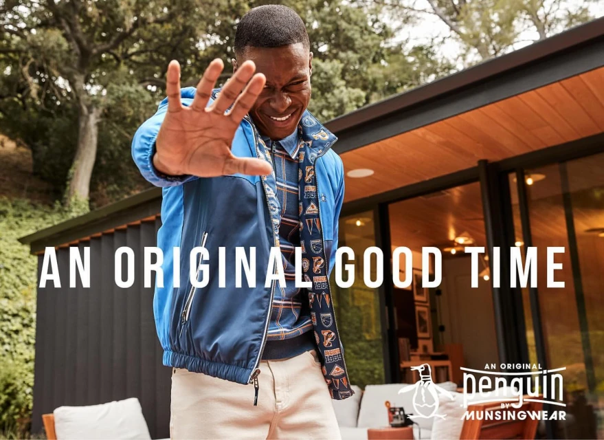 An African-American man smiles and puts his hand up in front of a building, wearing a blue jacket and khaki pants for a fashion advertisement.