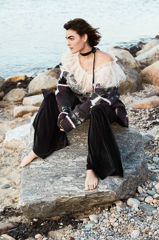 Model seated on a rocky shore wearing an off-shoulder frilled top and dark velvet trousers, photographed by Ben Watts.