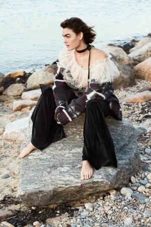 Model seated on a rocky shore wearing an off-shoulder frilled top and dark velvet trousers, photographed by Ben Watts.
