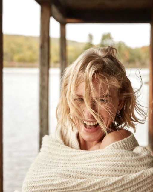 Smiling woman with blonde hair wrapped in a cream-colored knit blanket on a porch overlooking a lake and trees in a daylight setting.