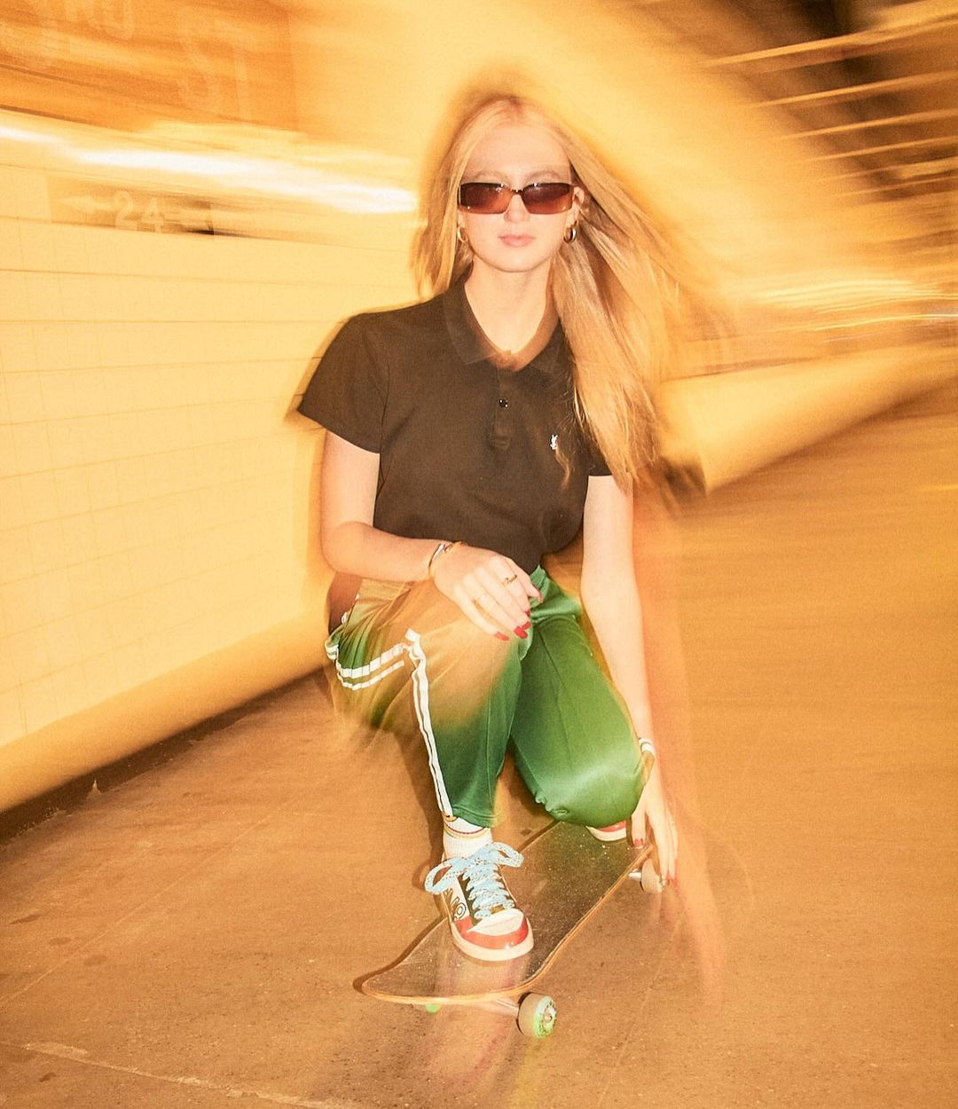 Young woman with long blonde hair skateboarding in an indoor urban location, wearing sunglasses, black shirt, and green satin pants.