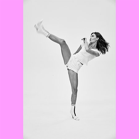 A woman in white shorts and a t-shirt is doing a martial arts kick with one leg extended high and the other leg on the floor.
