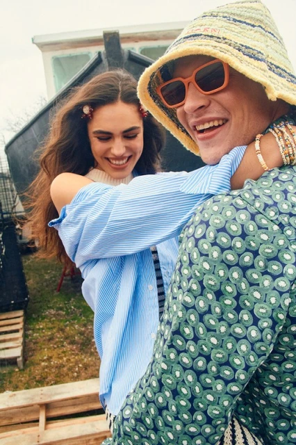 A smiling young woman hugs a smiling young man wearing a patterned shirt, hat, and orange glasses in an outdoor setting with a boat.