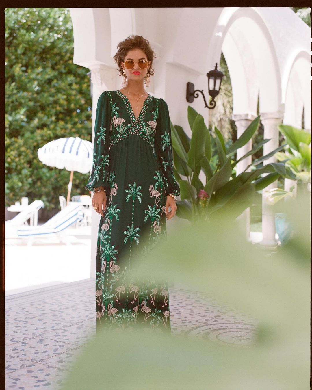 Woman in a green dress with flamingo and palm tree patterns standing on a patio with white arches and tropical plants.