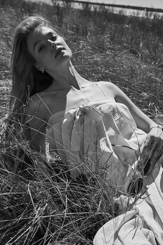 Black and white portrait of a blonde woman lying in tall grass outdoors, captured by Ben Watts.