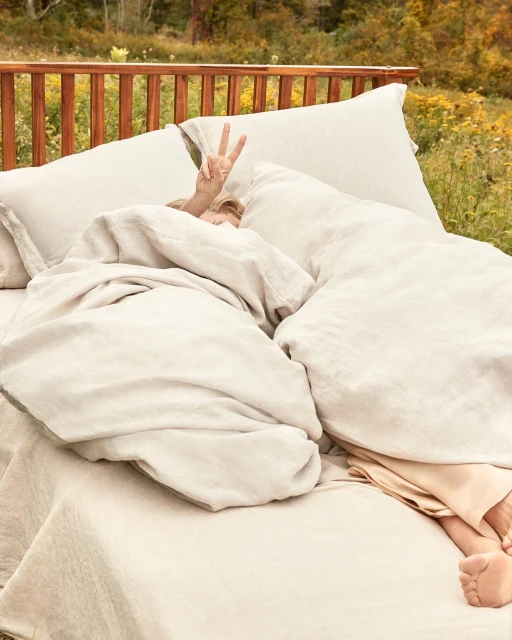 Person in bed outdoors gives a peace sign. Comfortable bed with white linen bedding is set up in a grassy, wooded area.