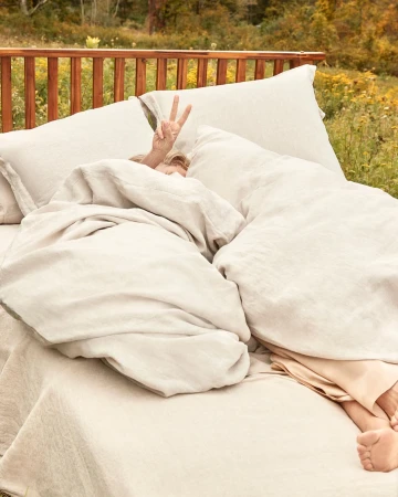 Person in bed outdoors gives a peace sign. Comfortable bed with white linen bedding is set up in a grassy, wooded area.