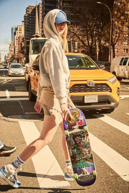 Young woman crosses city street with skateboard, wearing a hoodie and hat. Yellow taxi and buildings are in the background.