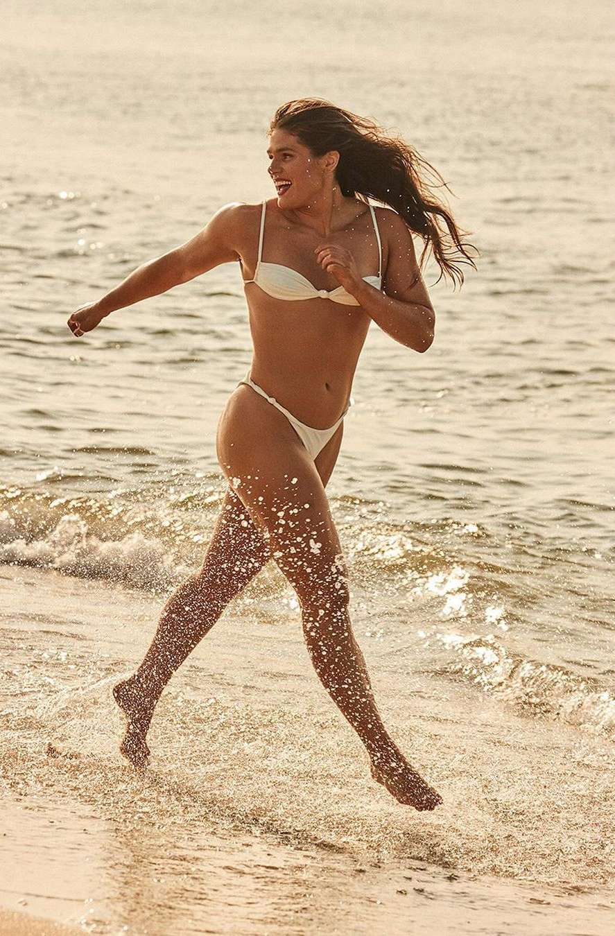A woman in a white bikini running through shallow water on a sandy beach, captured in warm sunlight.