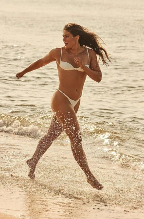 A woman in a white bikini running through shallow water on a sandy beach, captured in warm sunlight.