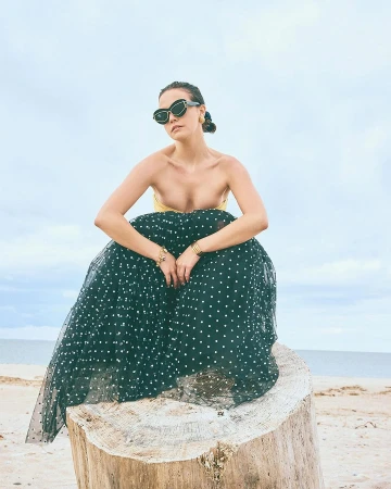 Woman wearing black sunglasses and a polka dot tulle skirt crouched on a tree stump by the ocean, photographed by Ben Watts.