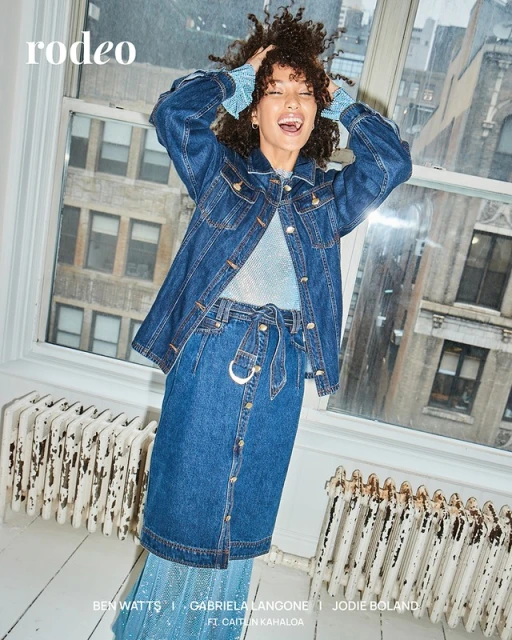 A woman smiles while posing indoors in a denim jacket, skirt, and shiny top. The shot is framed by a window and radiator.