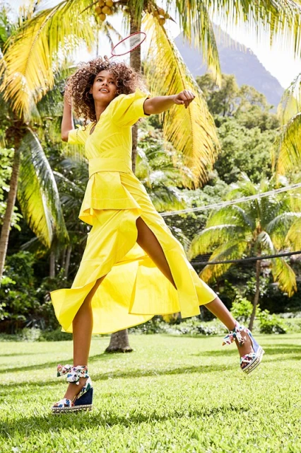 A woman in a yellow dress plays badminton on a grassy lawn with palm trees, showcasing a bright and active outdoor scene.