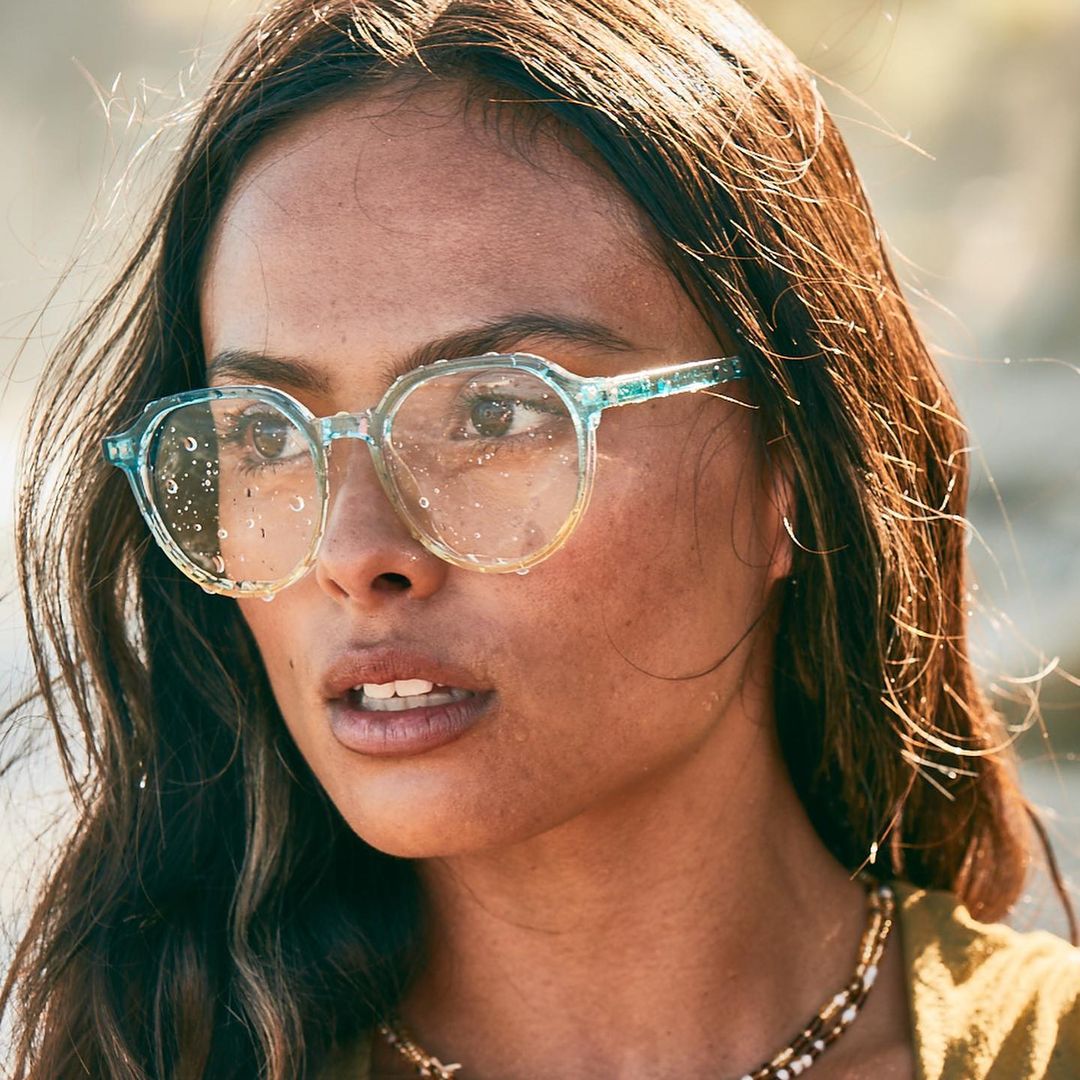 Close-up portrait of a woman wearing eyeglasses with water droplets on the lenses, with a blurred outdoor background in soft lighting.