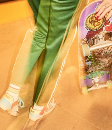 A person in green pants, wearing colorful sneakers, stands with a skateboard featuring graphics of art, coins, and stickers.