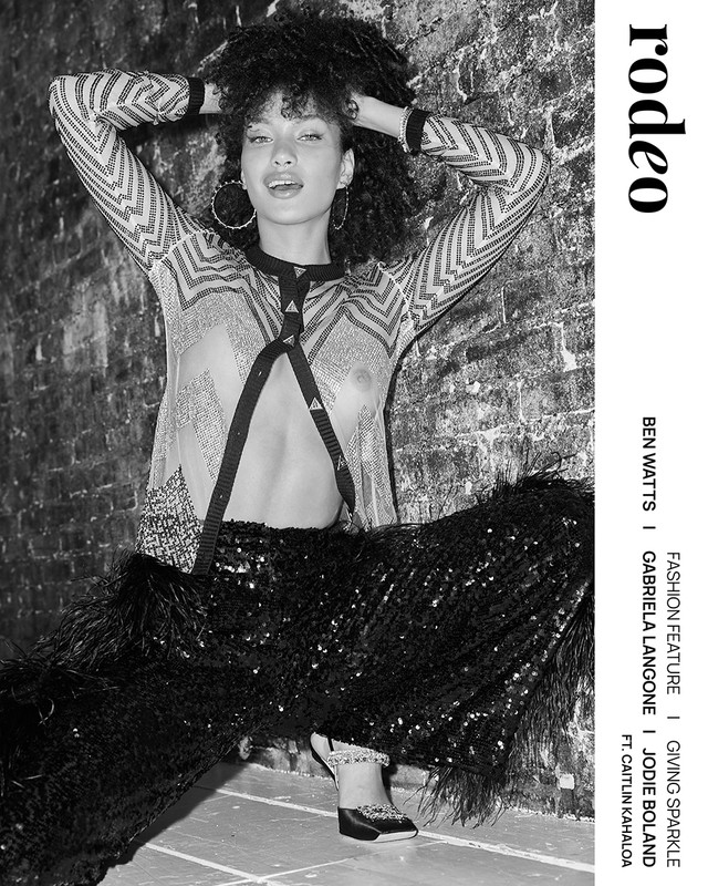 Black-and-white fashion shot of a model with curly hair posing in a patterned top, sequined pants with feathered trim, and platform shoes.