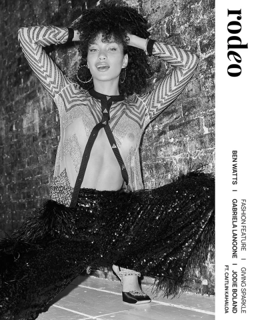 Black-and-white fashion shot of a model with curly hair posing in a patterned top, sequined pants with feathered trim, and platform shoes.