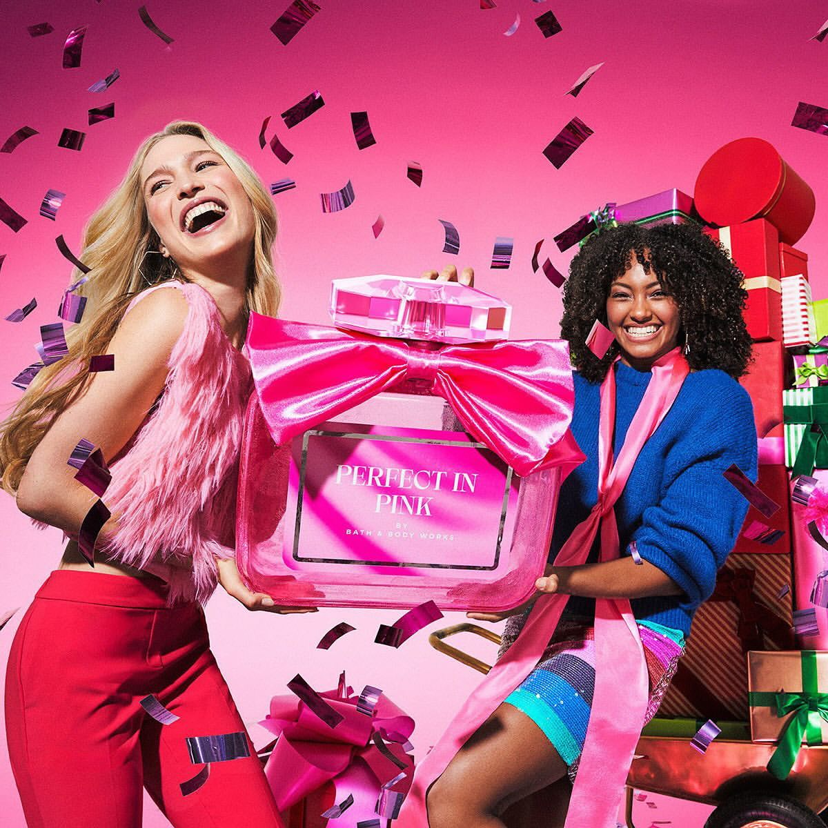 Two smiling women hold a large pink perfume bottle with a bow in a studio setting with confetti and stacked gifts against a pink backdrop.