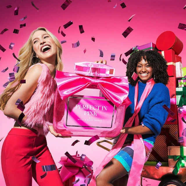 Two smiling women hold a large pink perfume bottle with a bow in a studio setting with confetti and stacked gifts against a pink backdrop.