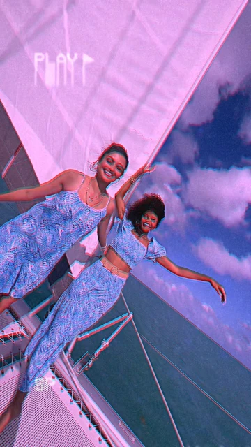 Two smiling women pose in matching blue patterned outfits on a sailboat under a cloudy sky, with a retro video filter overlay.