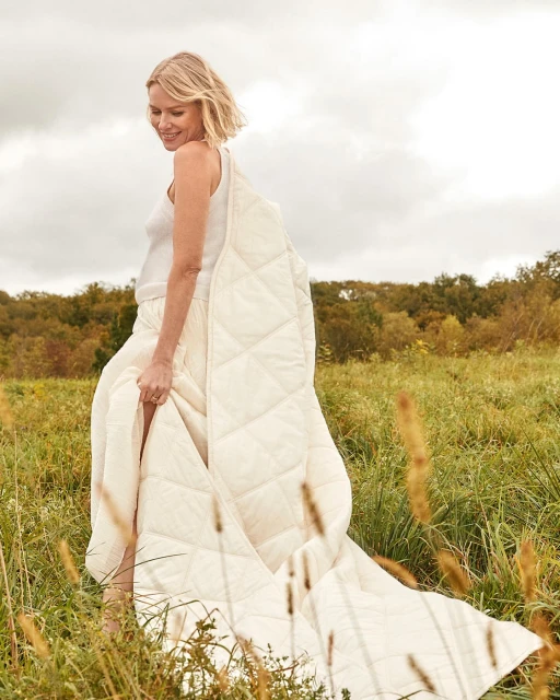 Adult woman wearing a white top and cream-colored skirt standing in a grassy field with a quilted blanket draped around her shoulders.