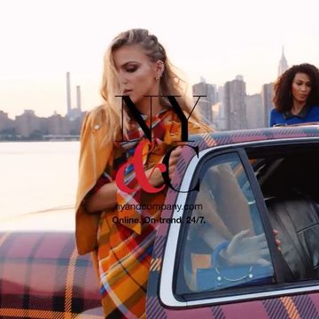 Woman in yellow jacket and plaid blanket near a car with a city skyline in the background during golden hour.