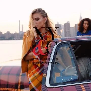 Woman in yellow jacket and plaid blanket near a car with a city skyline in the background during golden hour.