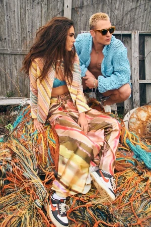 Two fashion models pose outdoors in colorful outfits and shoes, sitting atop tangled ropes with a wooden fence backdrop.