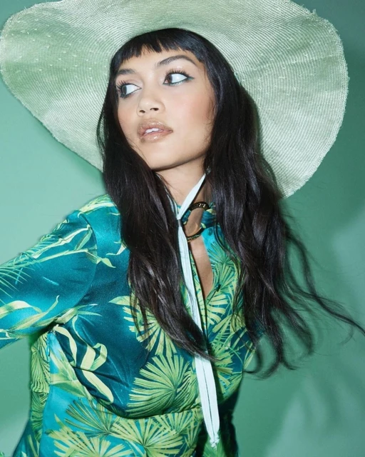 Portrait of a woman wearing a wide-brimmed hat and green tropical print dress. She is looking to the side against a green background.