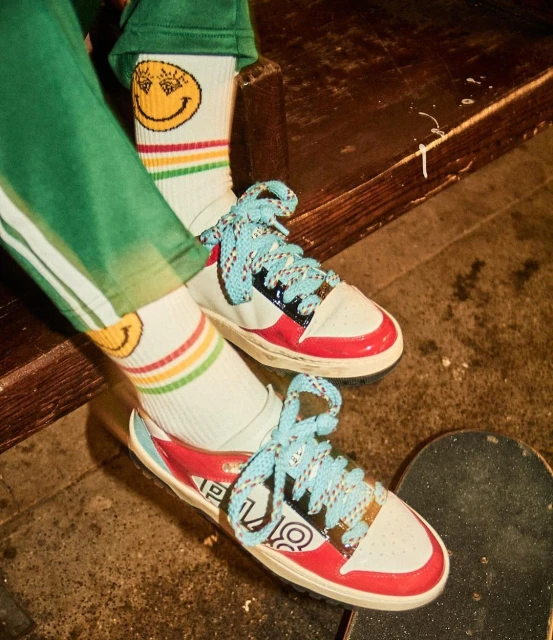 Close-up of a pair of sneakers with turquoise laces on a skateboard, styled with smiley-face socks and green striped pants.