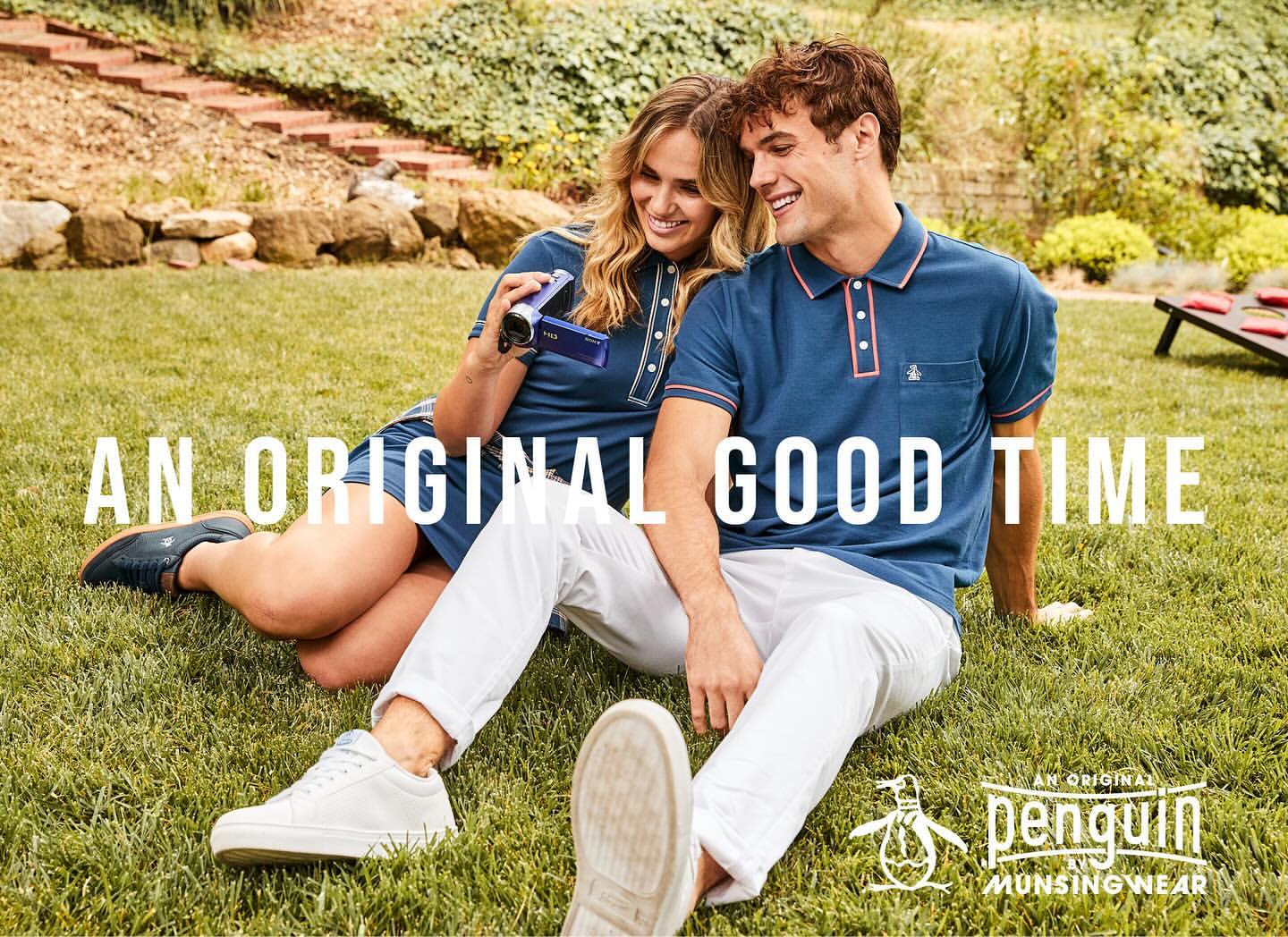 A smiling couple in matching polo shirts sits on a lawn outdoors; the woman holds a vintage camcorder, and the man wears white pants.