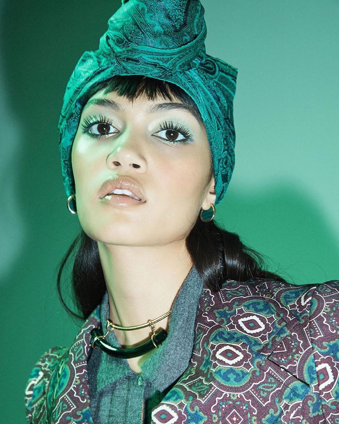 Portrait of an adult woman with a green paisley headscarf, heavy makeup, and a patterned jacket against a light green background.