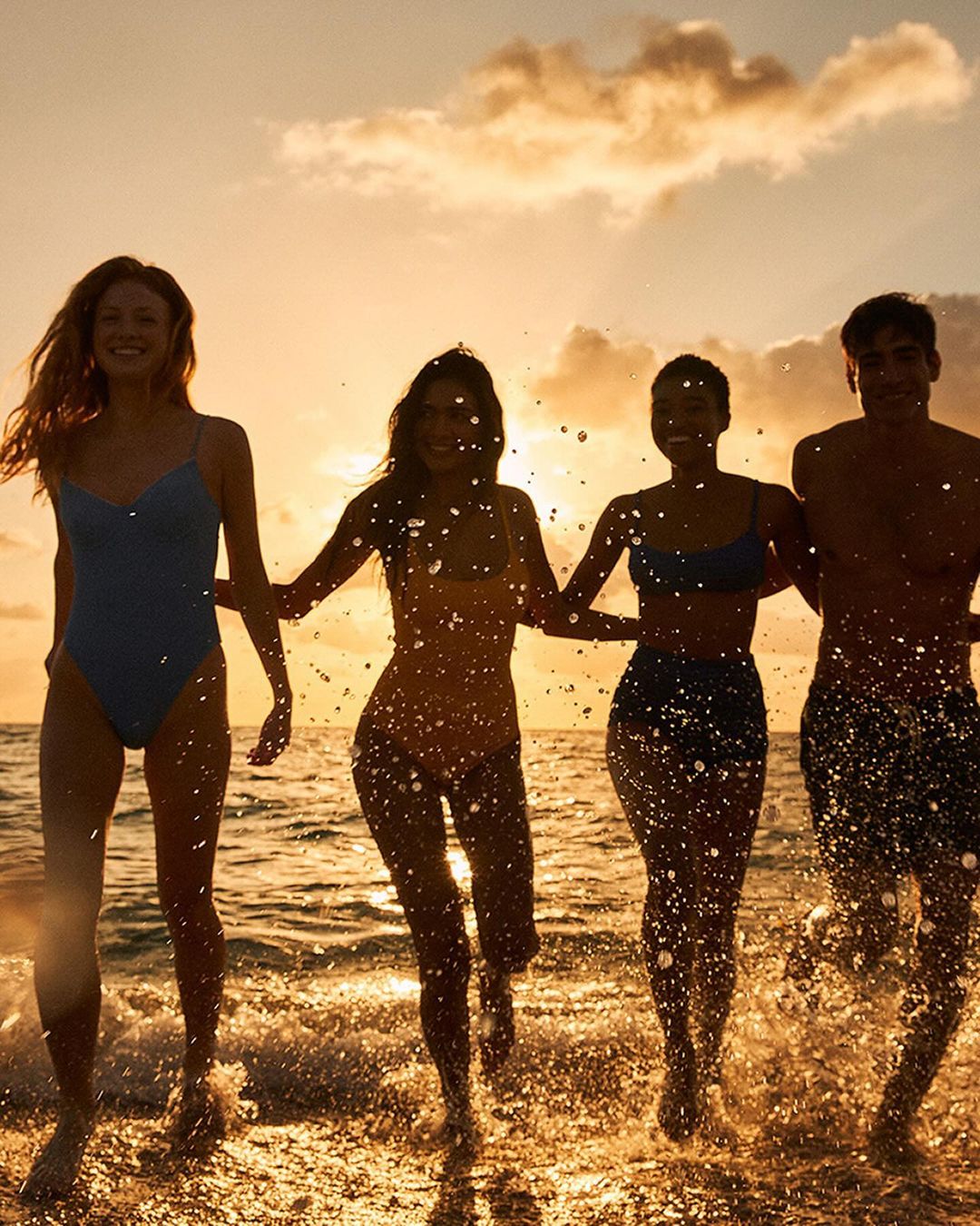 Four young adults in swimsuits walk along a beach at sunset. Splashes of water highlight the figures against a golden sky.