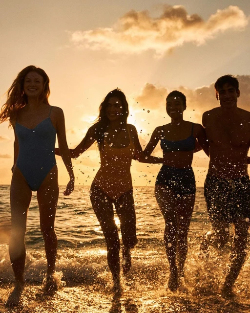 Four young adults in swimsuits walk along a beach at sunset. Splashes of water highlight the figures against a golden sky.
