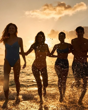 Four young adults in swimsuits walk along a beach at sunset. Splashes of water highlight the figures against a golden sky.