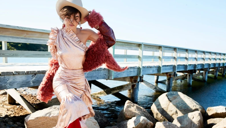 Woman in shiny one-shoulder dress and cowboy hat wearing a fluffy pink coat walking near a wooden dock